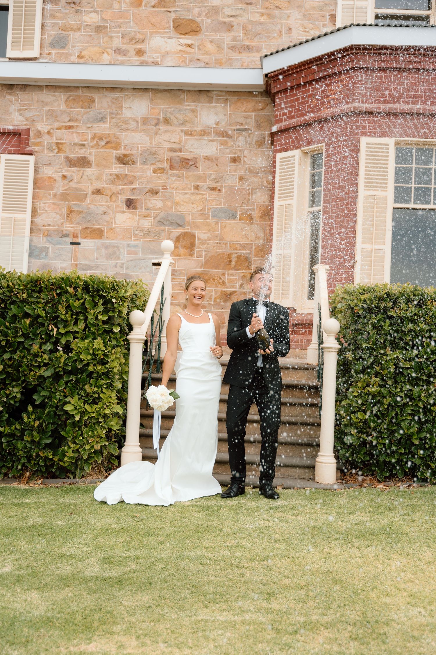Bride and groom popping champagne bottle on lawn outside stone building wedding venue.