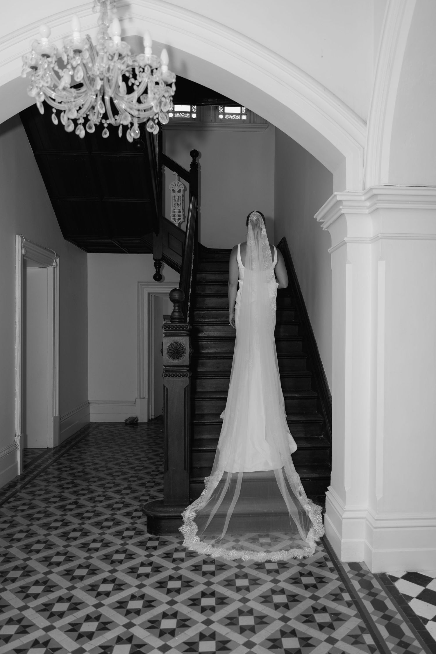 Black and white photo of bride in long veil standing by grand staircase with chandelier.