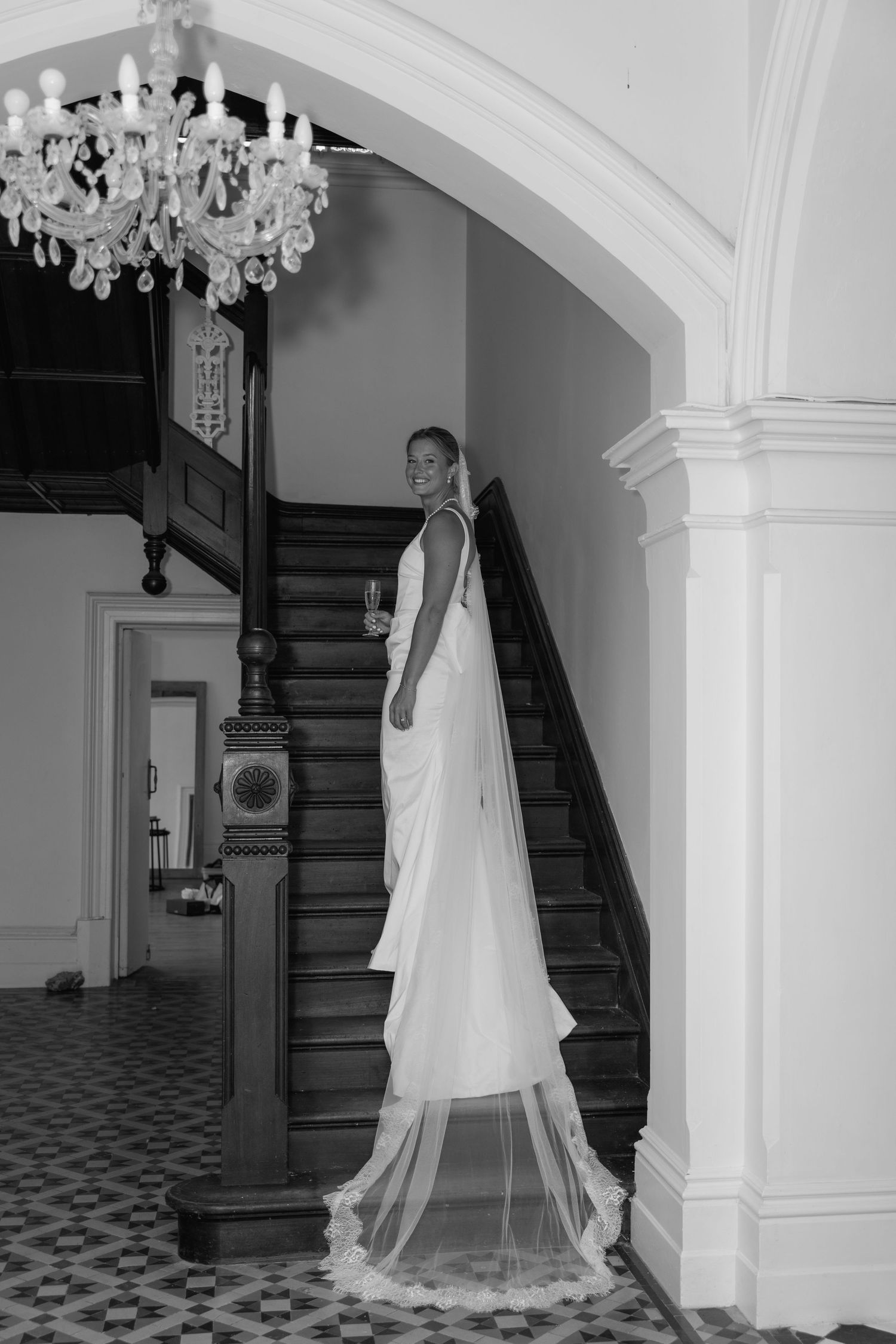 Bride in elegant white gown with long veil stands on staircase beneath chandelier in black and white photo.