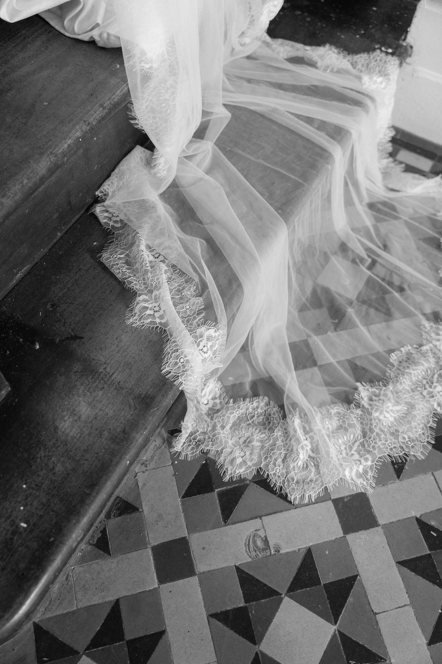 Black and white close-up of a bridal veil train with lace trim on tiled church floor.