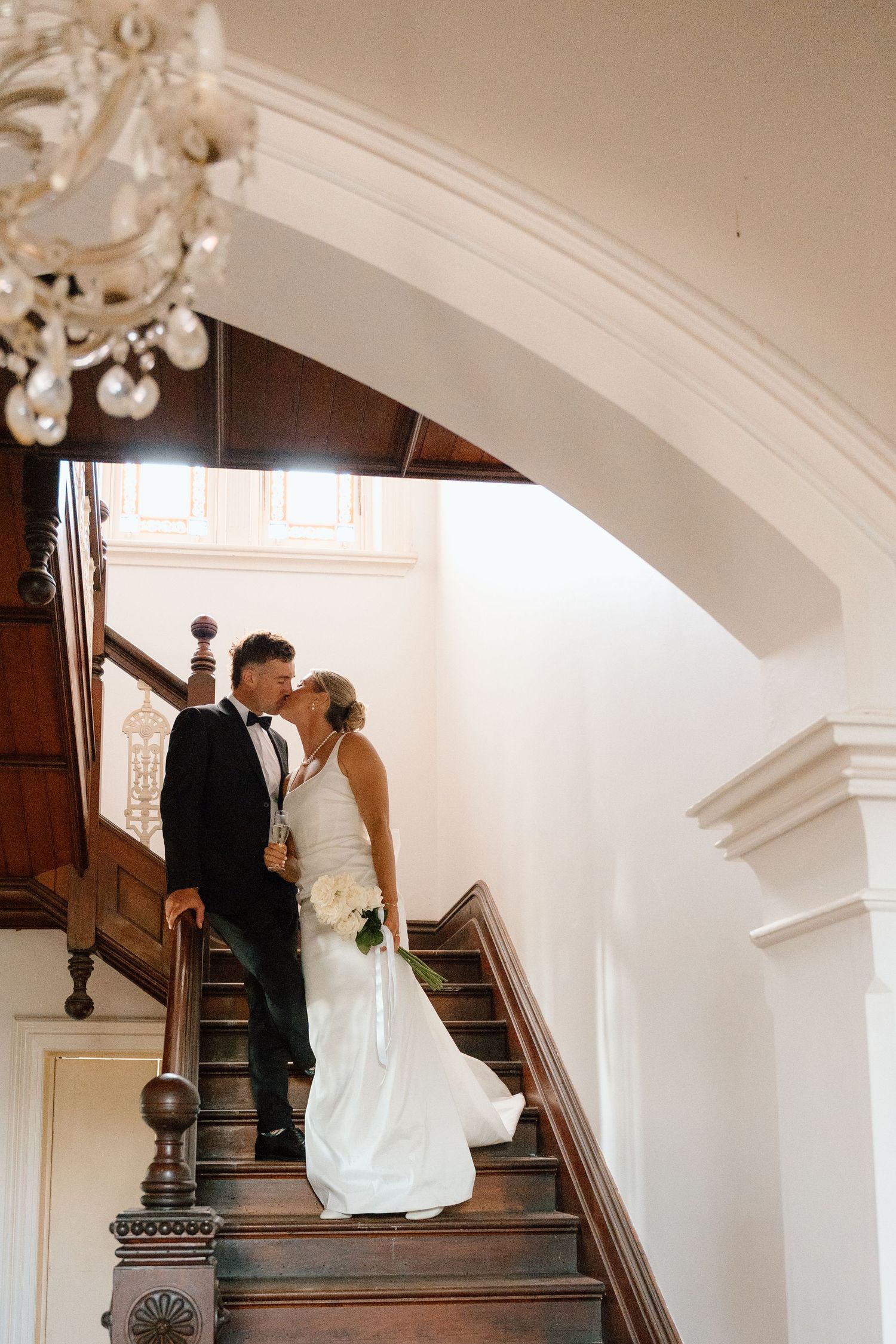 Bride and groom sharing a kiss on elegant wooden staircase beneath a crystal chandelier.