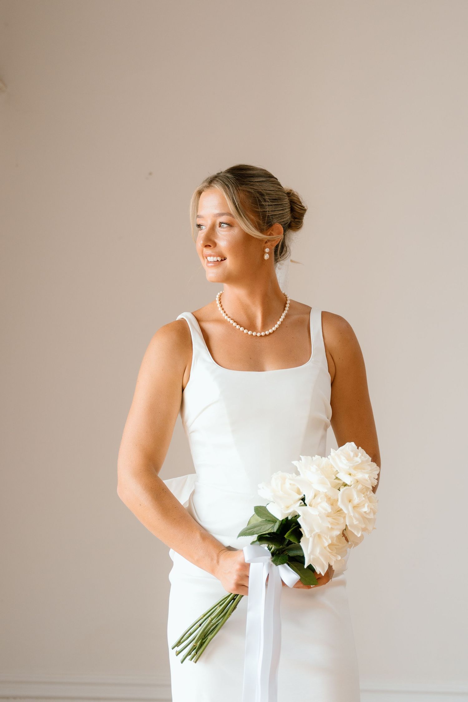 Smiling bride in white square-neck gown holding white hydrangea bouquet with pearl necklace.