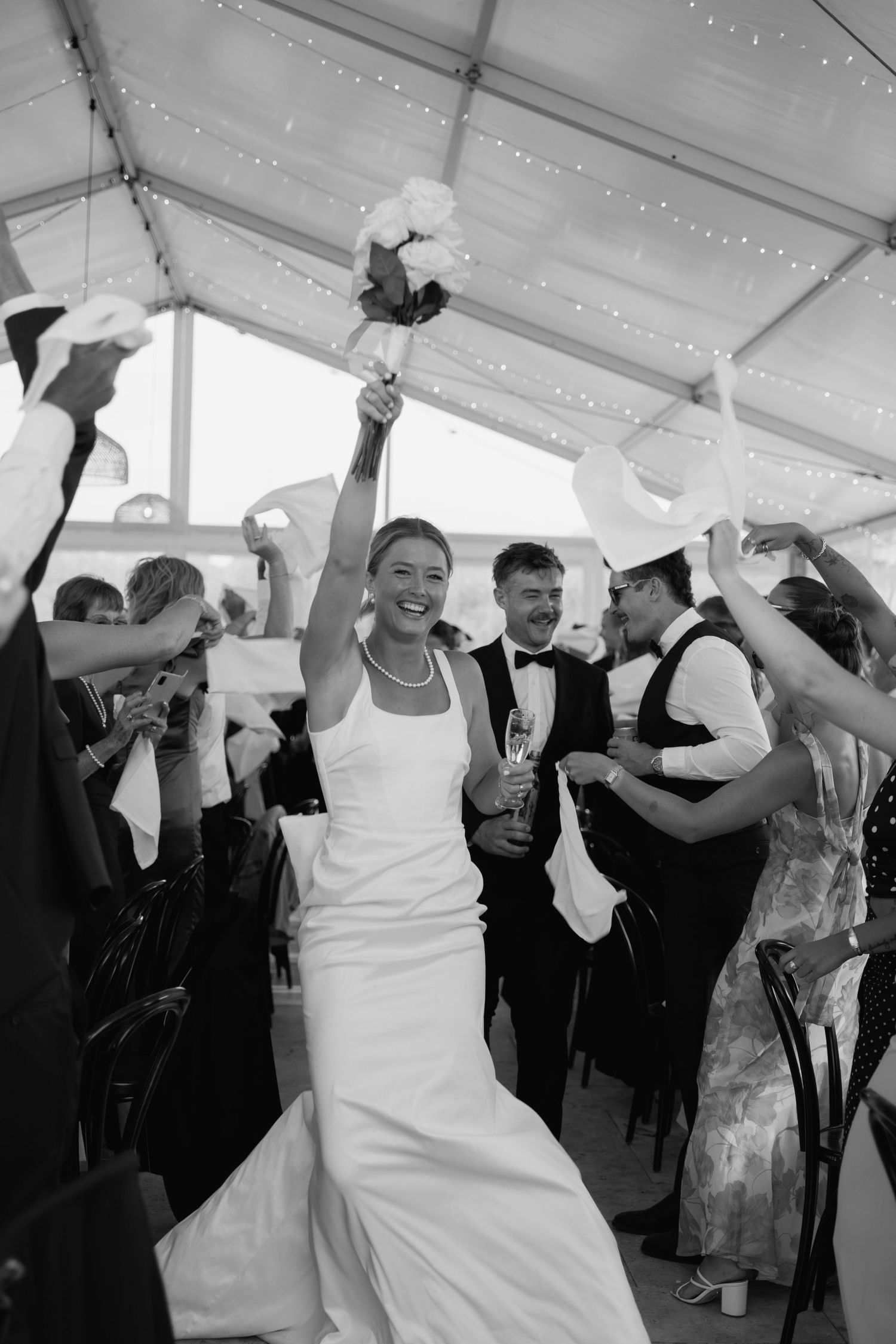 Bride raises bouquet joyfully while walking aisle with groom at elegant tented wedding reception.