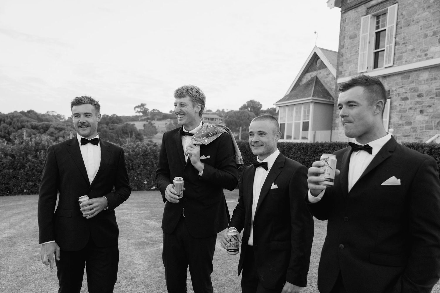 Black and white photo of four groomsmen in tuxedos laughing outside historic stone building.