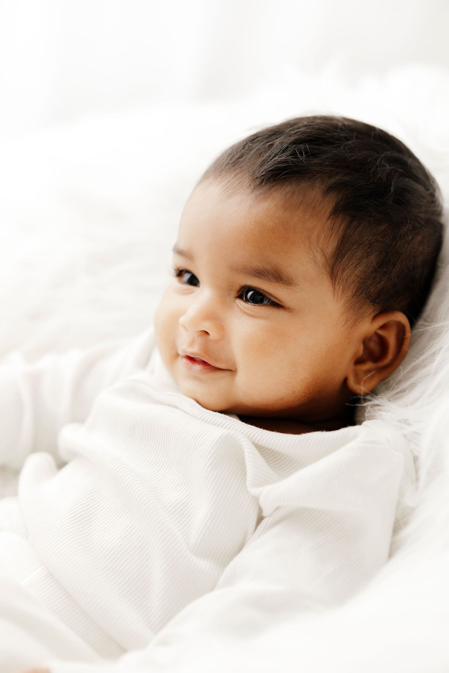 Baby girl in white lace dress and ruffle socks stands against white blocks, smiling in studio photo.