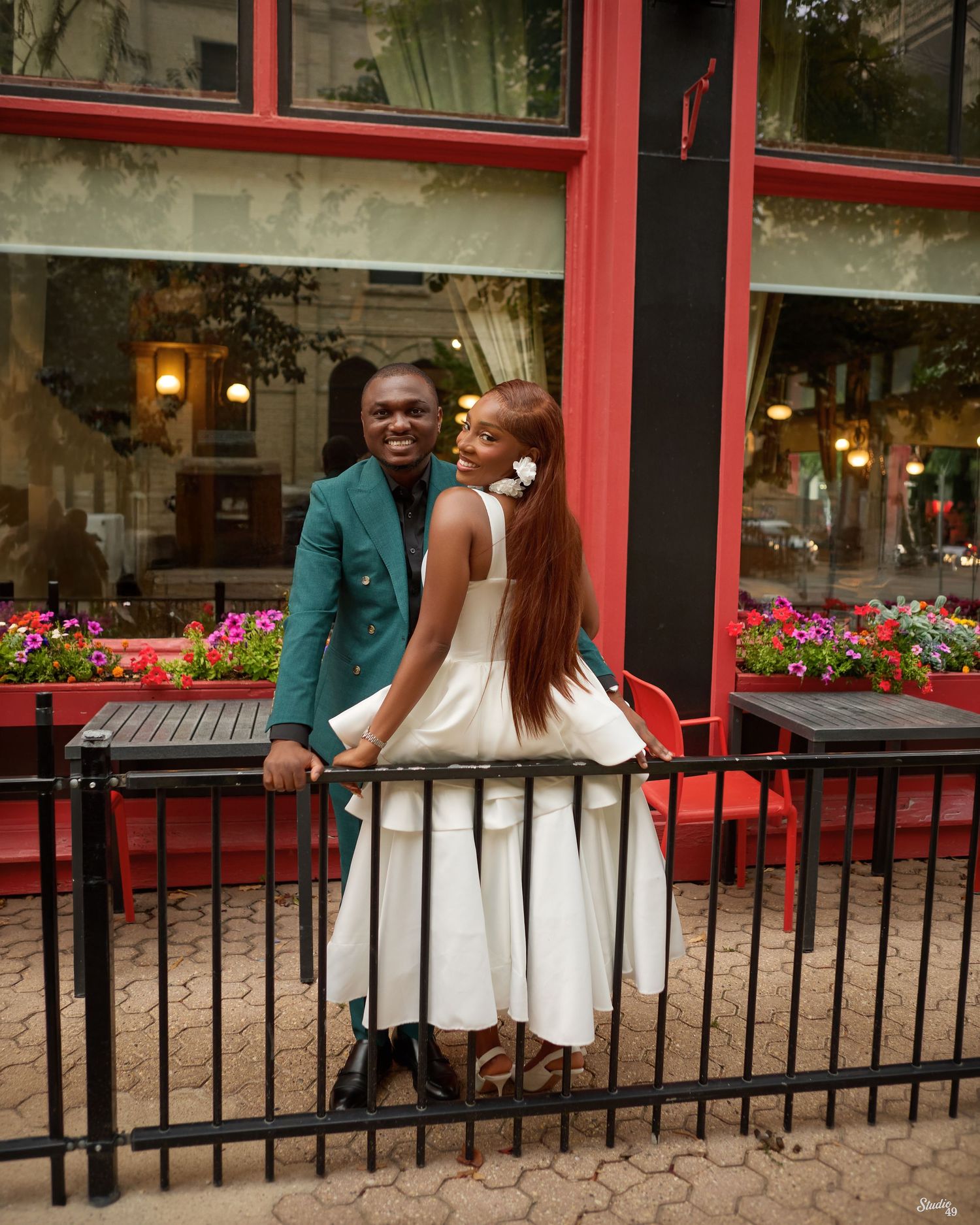 Couple posing outdoors at a restaurant patio, woman in white dress, man in green blazer.