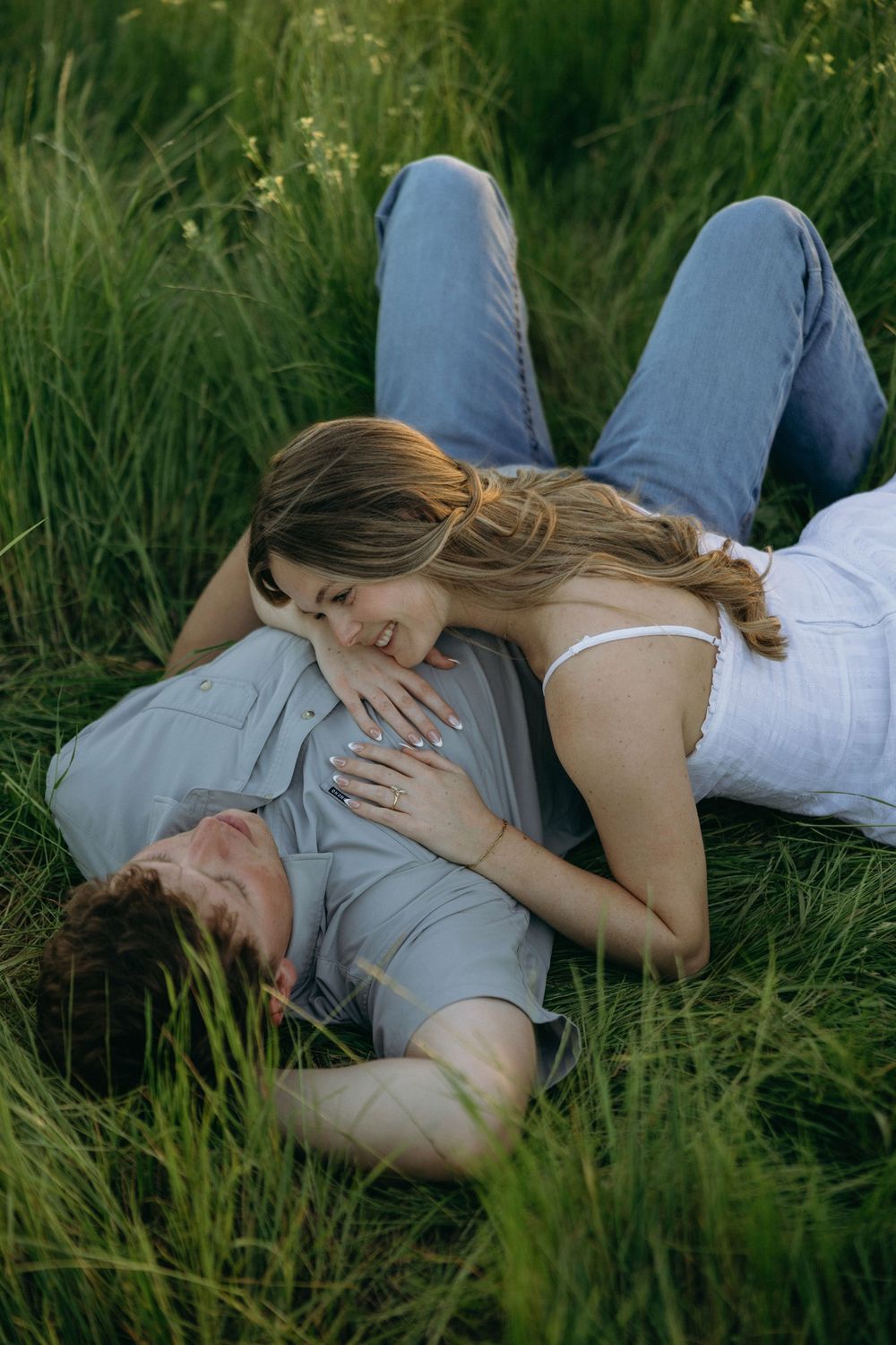 Couple lying in lush green grass, woman smiling and resting on mans chest during romantic outdoor photo session.