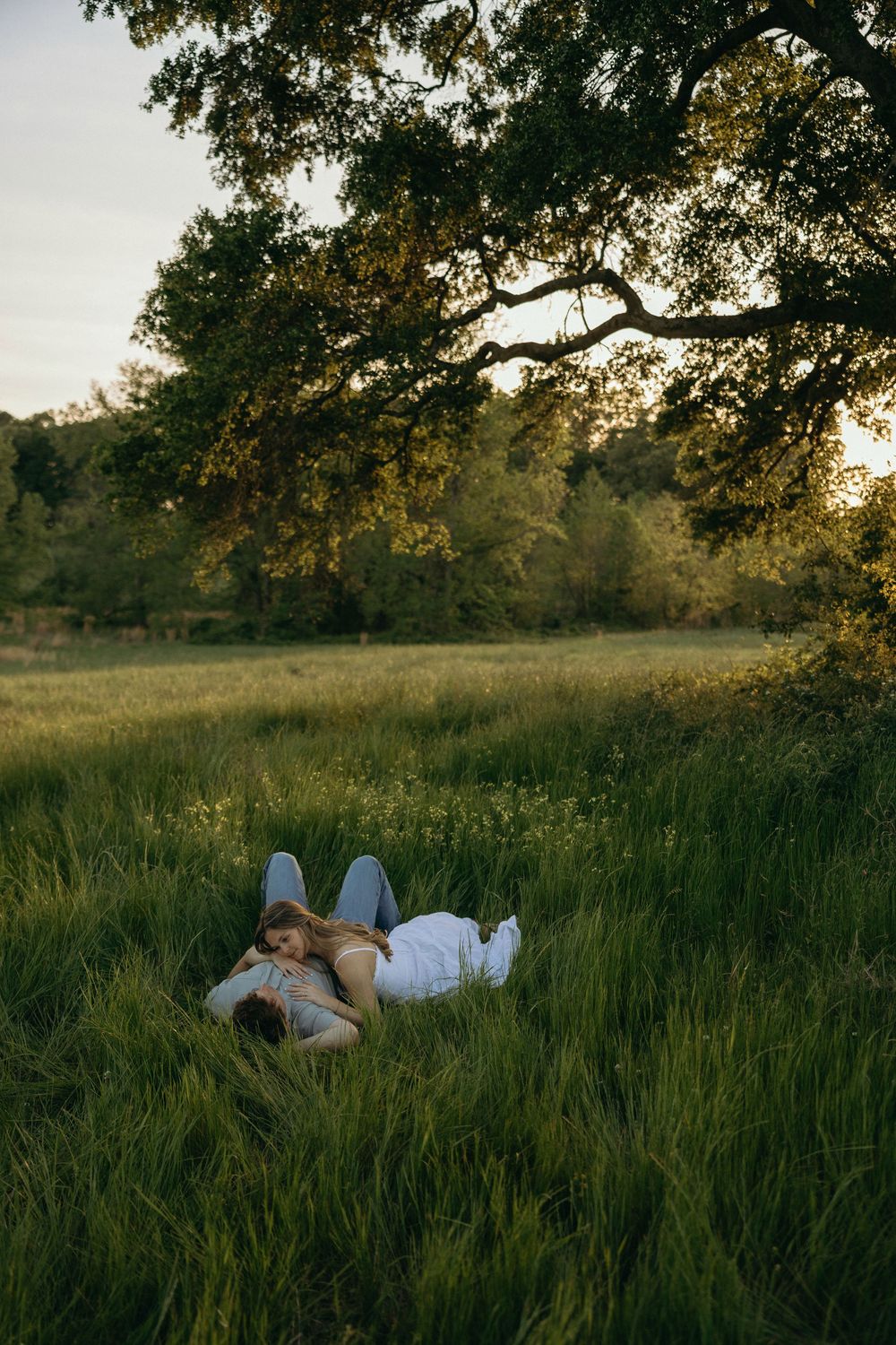 Couple lying in tall green grass beneath a large oak tree during thier engagement session south of Atlanta.
