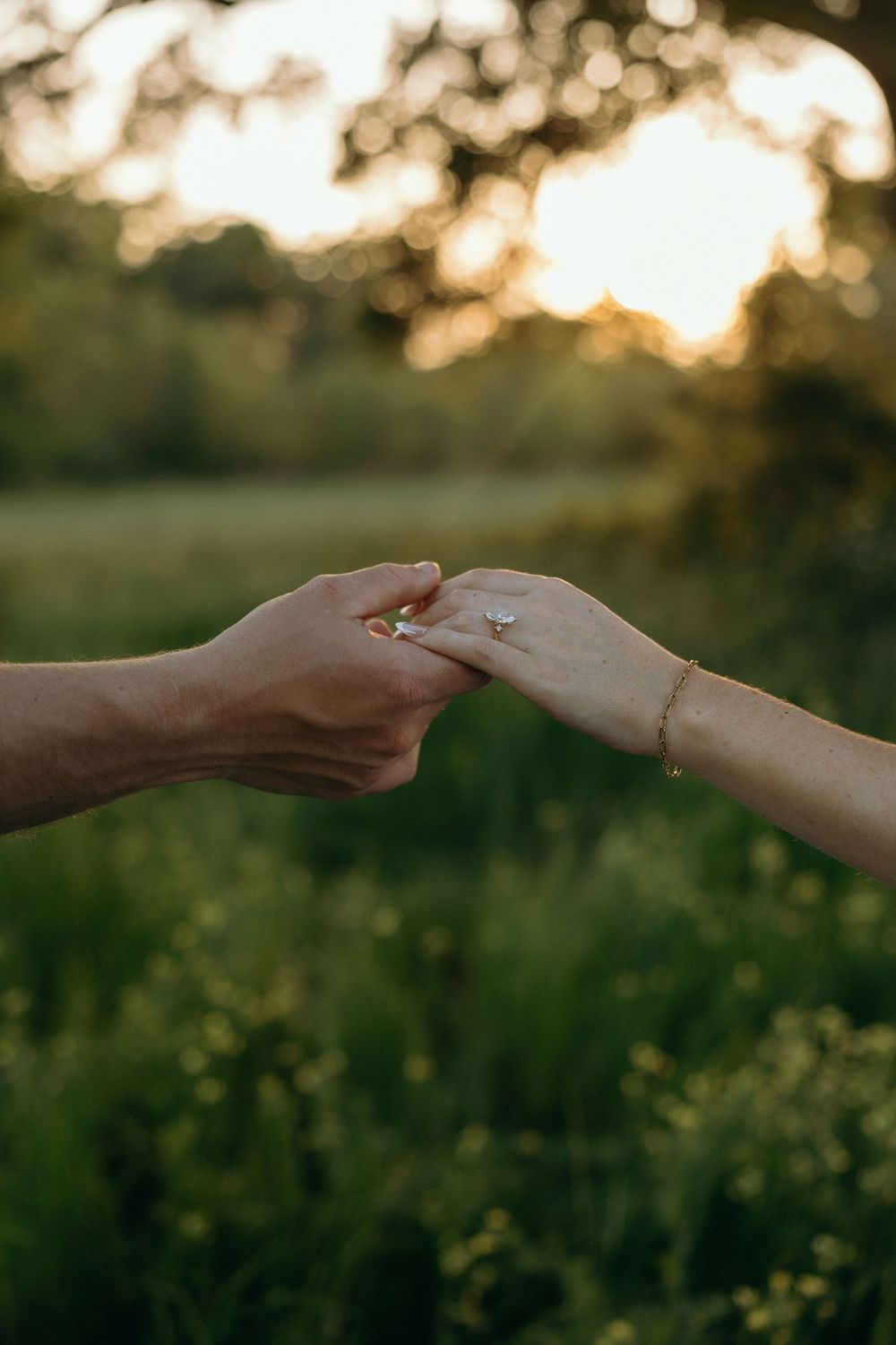 Two hands touching with an engagement ring at golden hour in a lush green field.