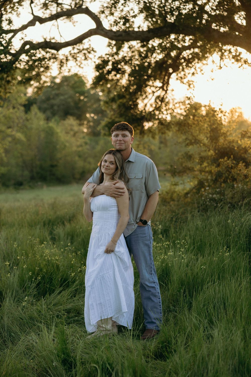 Couple embracing in lush green field at golden sunset, woman in white dress, man in gray shirt and jeans.