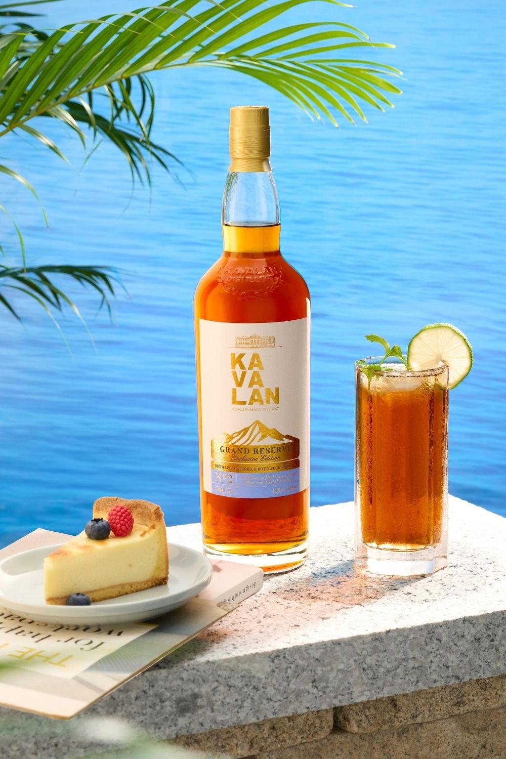 Kavalan Grand Remix whisky bottle beside a cocktail with lime and cheesecake, set against a tropical blue ocean backdrop.