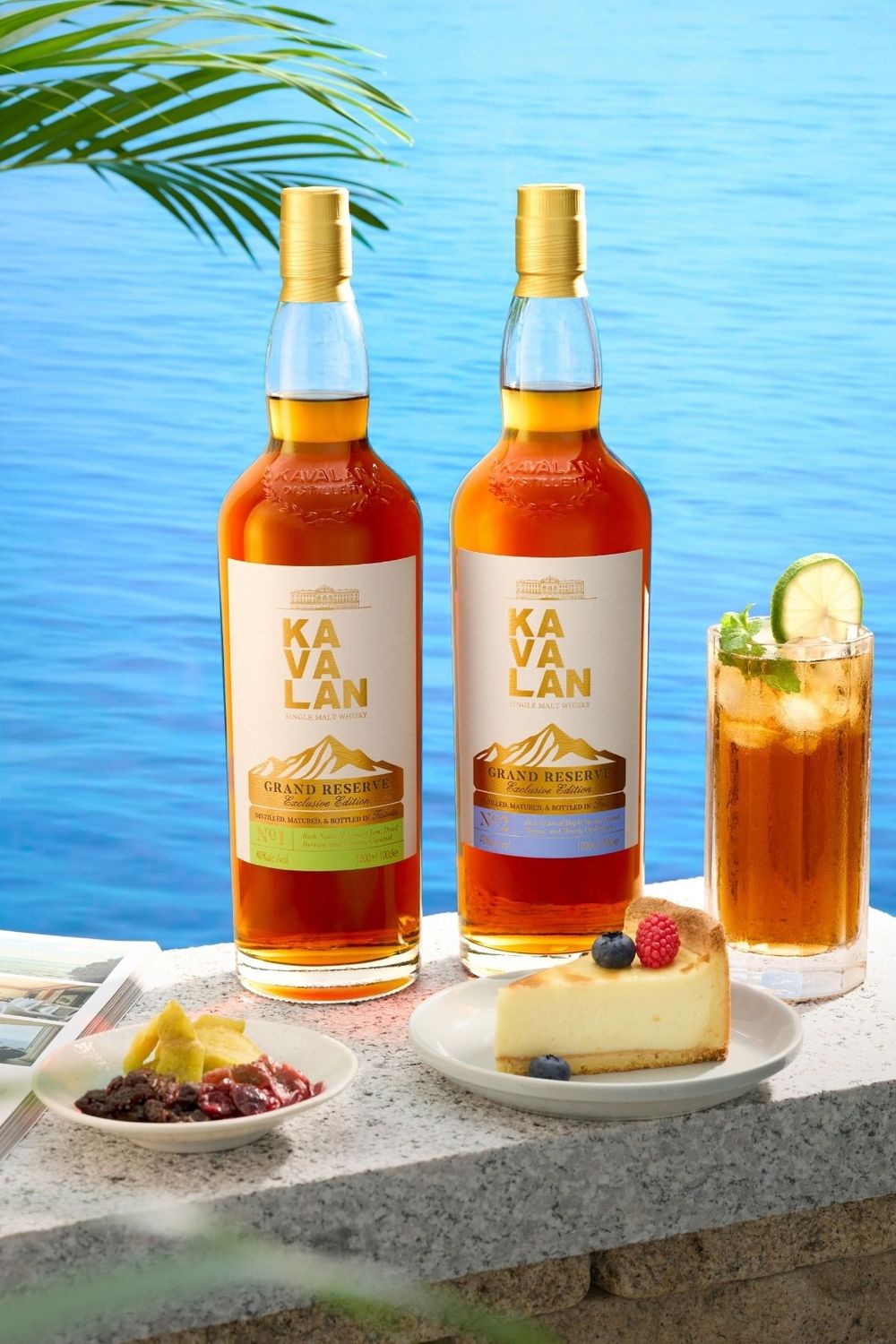 Two bottles of Kavalan Grand Reserve whisky displayed outdoors with cheesecake, tropical drink, and ocean view backdrop.