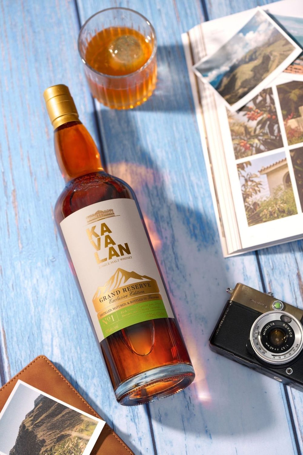 Kavalan Grand Reserve whisky bottle on blue wood surface with a glass of whisky, vintage camera, and travel photos.