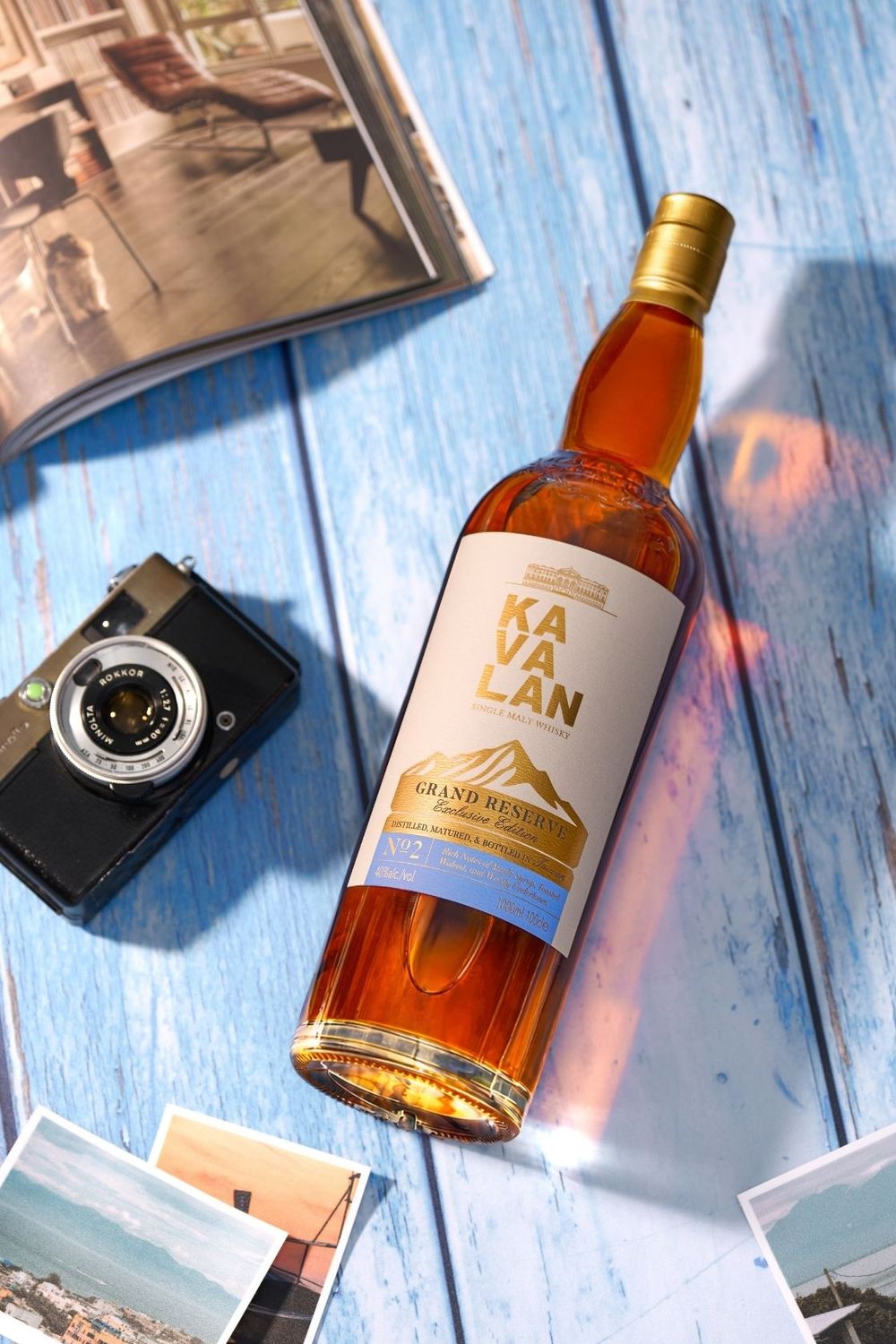 Kavalan Grand Reserve whisky bottle on blue wooden surface with vintage camera, travel photos, and magazine.