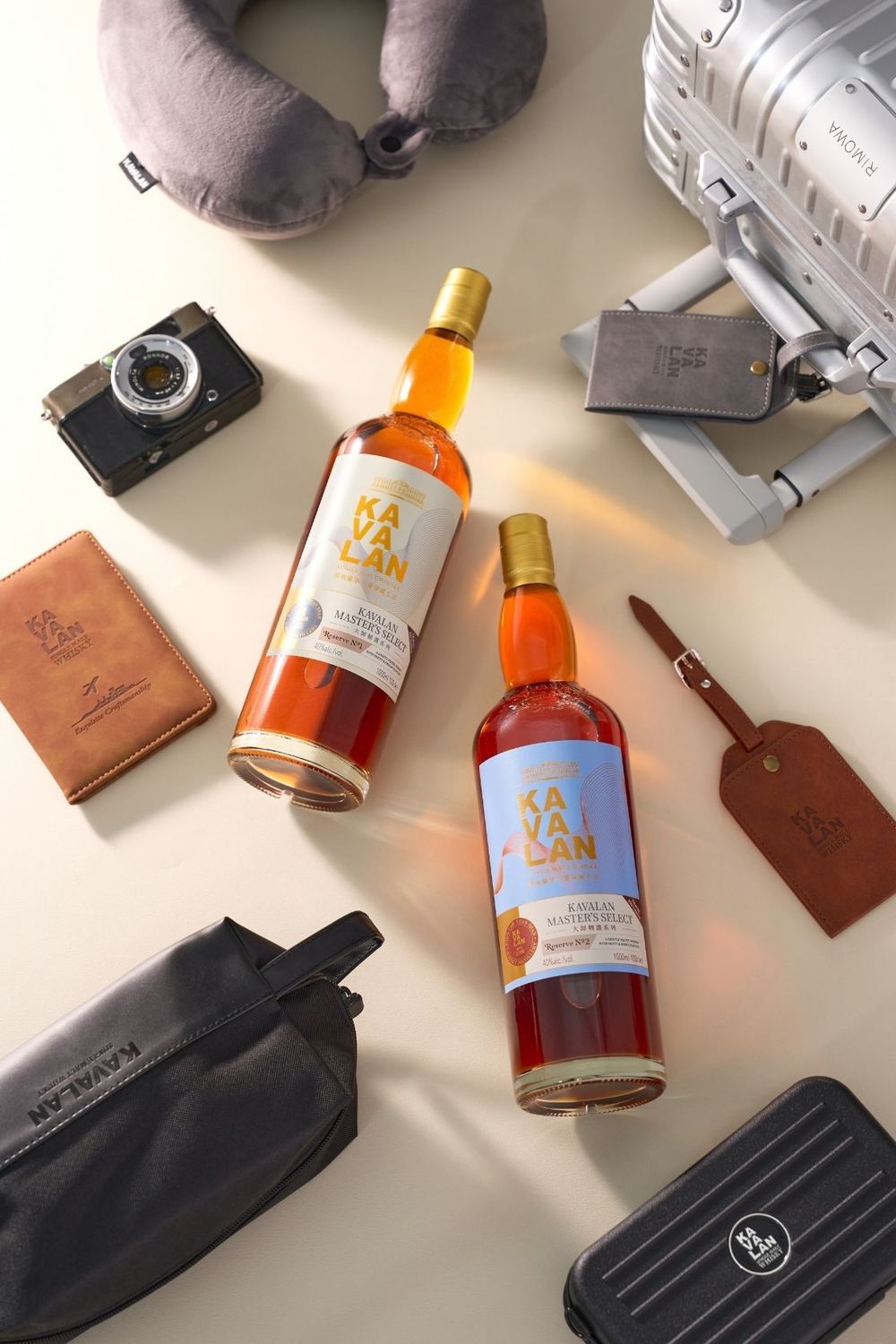 Two Kavalan whisky bottles displayed with travel accessories including a camera, luggage, passport holder, and neck pillow.