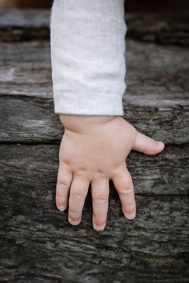 Close-up of a small child's hand resting on a weathered wooden log, wearing a white long-sleeve shirt.