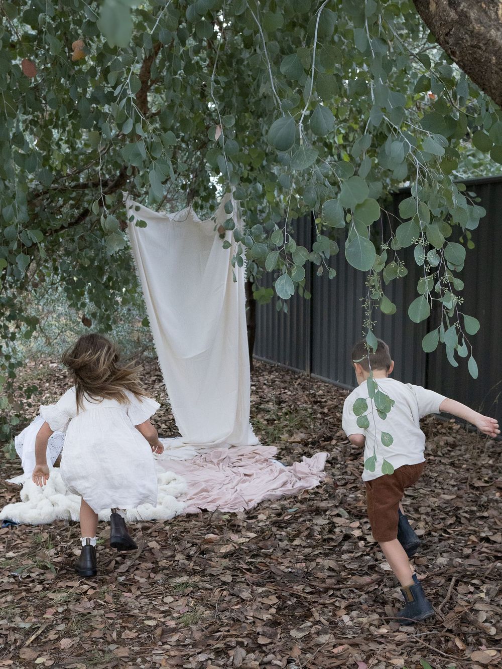 Two children playing outdoors under eucalyptus tree with white fabric hanging, surrounded by fallen leaves.