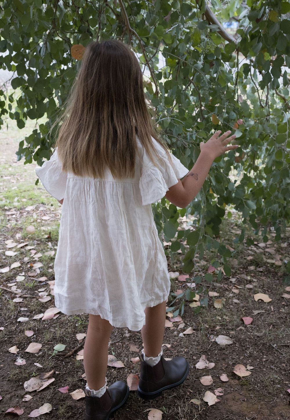 Young girl in white dress reaching for tree branches outdoors, wearing black shoes and lace socks.
