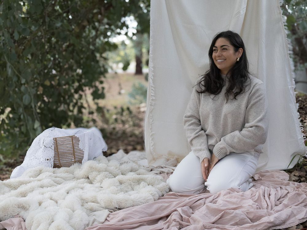 A smiling woman sits outdoors on pink fabric beside fluffy white wool, with a white curtain backdrop among trees.
