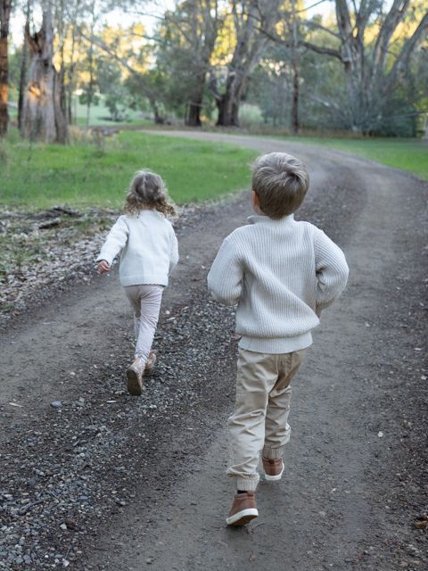 Two young children in white sweaters walk hand in hand down a tree-lined gravel path in a lush park.