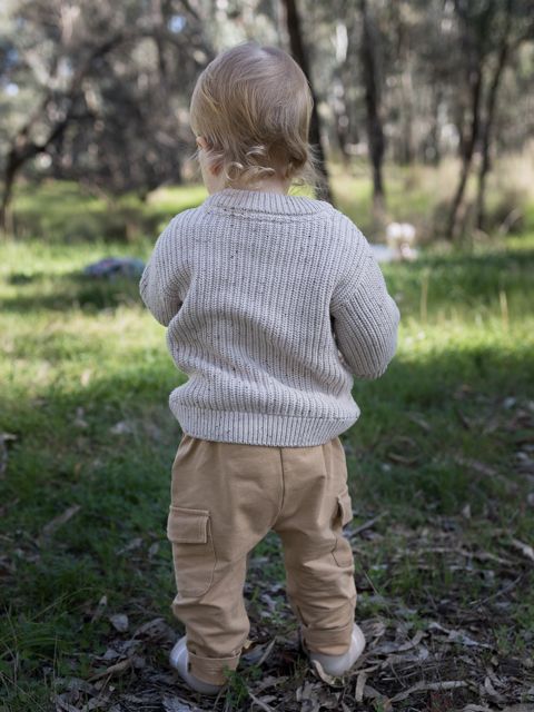 Toddler in beige knit sweater and tan cargo pants standing outdoors in a lush green garden, viewed from behind.