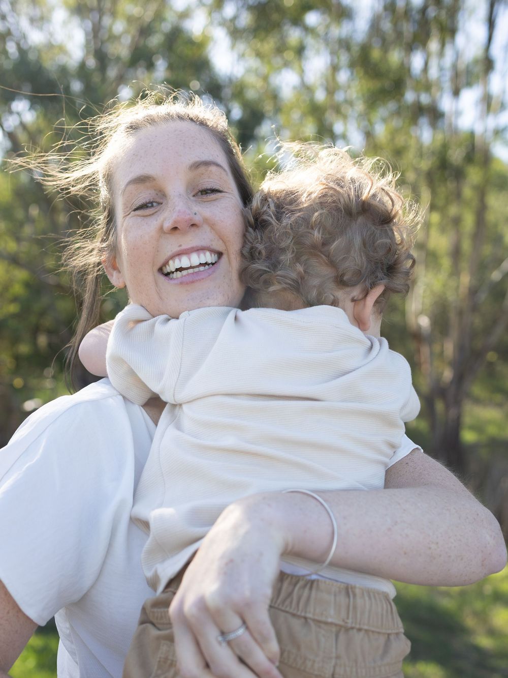 A smiling woman with freckles hugs a curly-haired toddler outdoors among green trees on a sunny day.