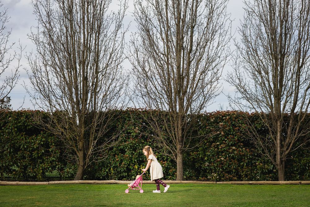 A mother walks with her young child in a green park, surrounded by bare winter trees and a lush hedge backdrop.