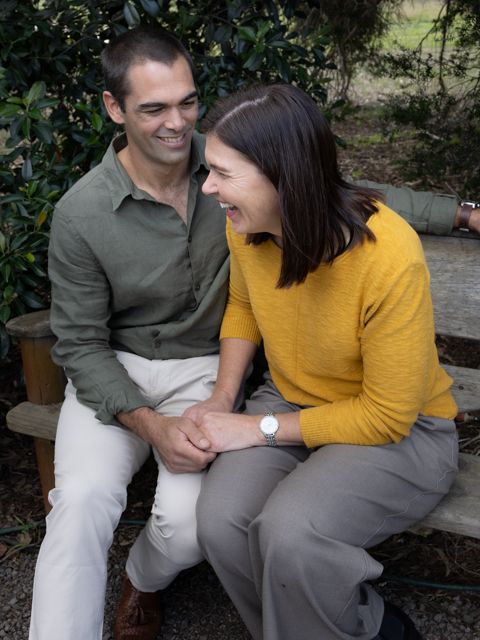 Happy couple sitting on a wooden bench outdoors, laughing together, wearing casual green and yellow sweaters.
