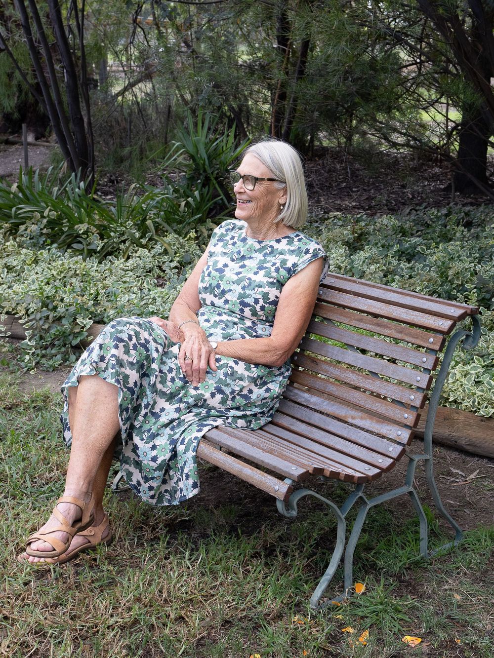 Older woman in floral dress sitting on wooden park bench surrounded by lush green garden foliage.