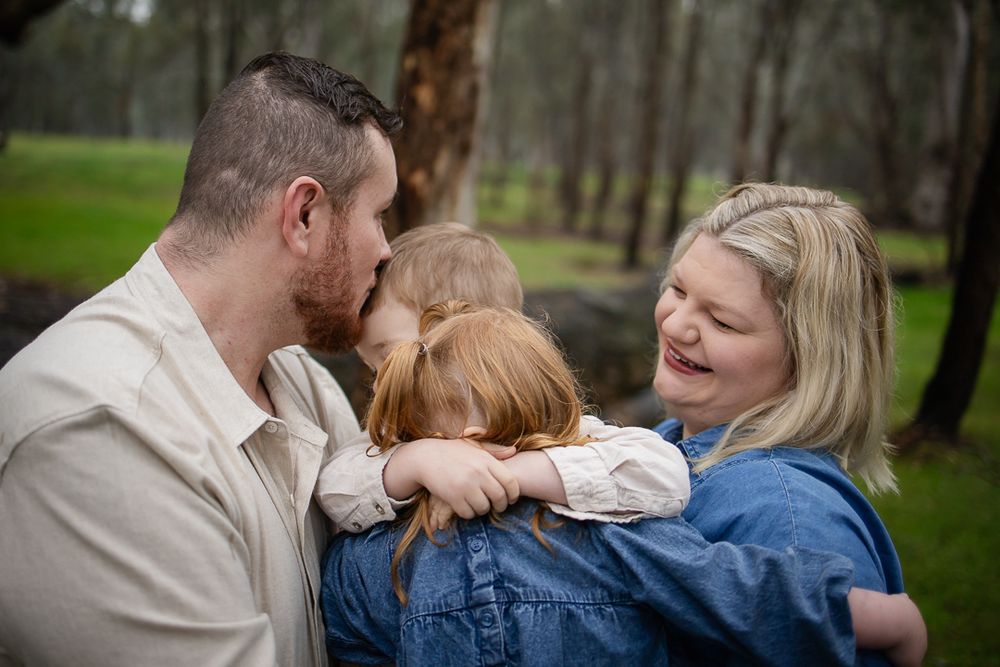 A happy family of three sharing a warm embrace outdoors among trees in a lush green park.