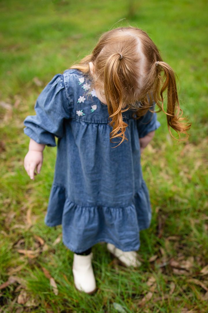 Young girl with red pigtails wearing a blue denim dress and white boots walking on green grass.