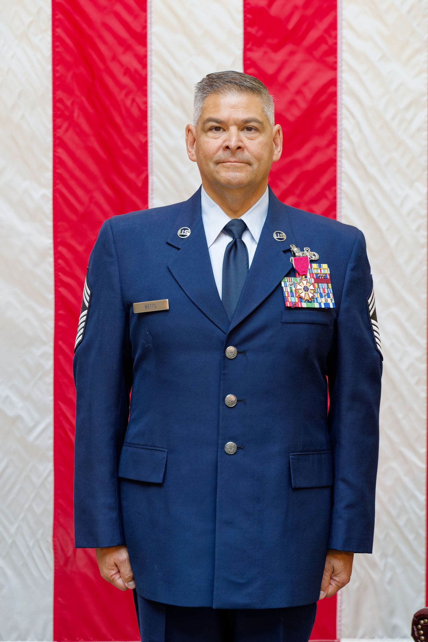 US Air Force senior non-commissioned officer in dress blue uniform with medals standing before American flag.