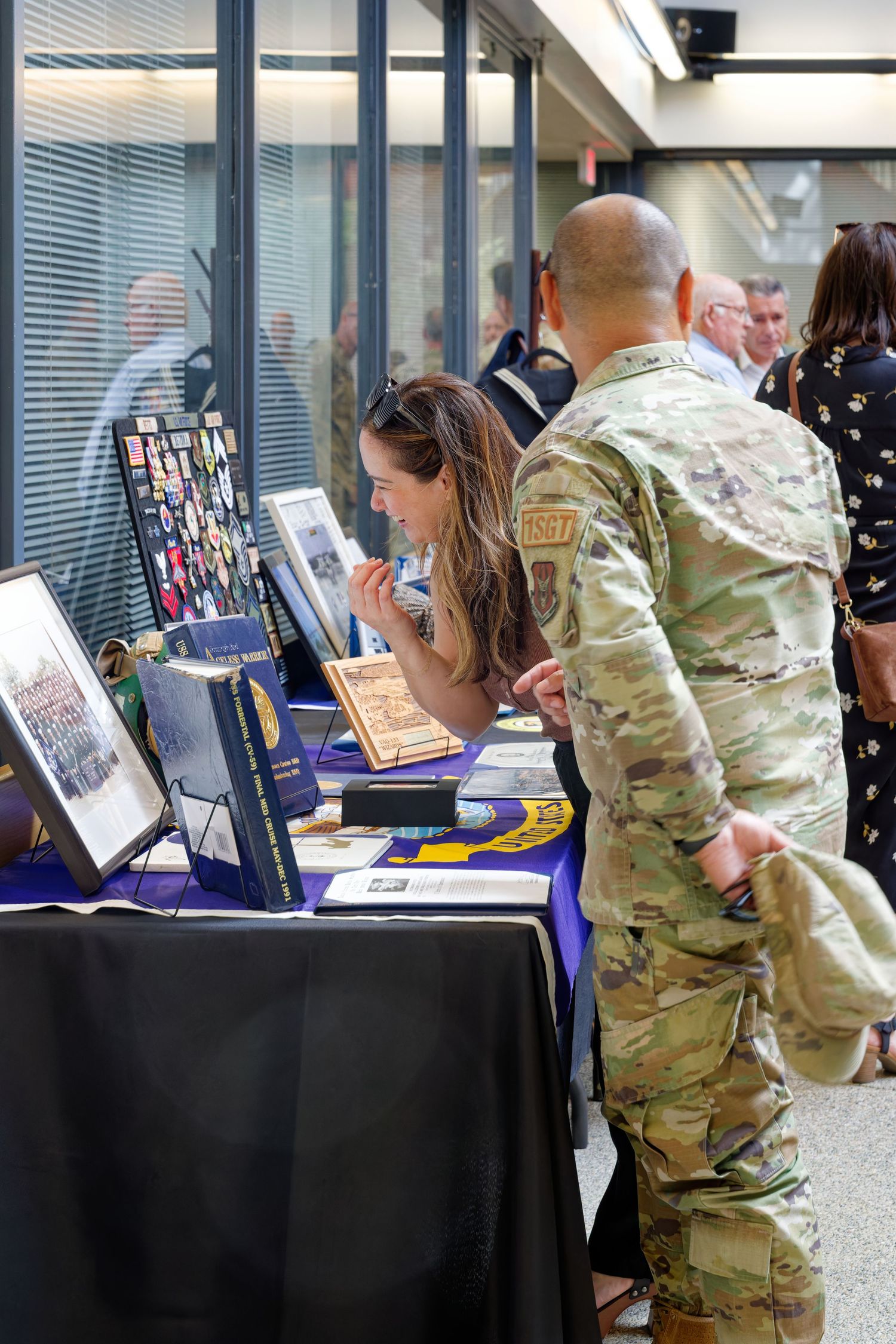 Soldier in camouflage uniform and woman examining military memorabilia at an indoor display table.