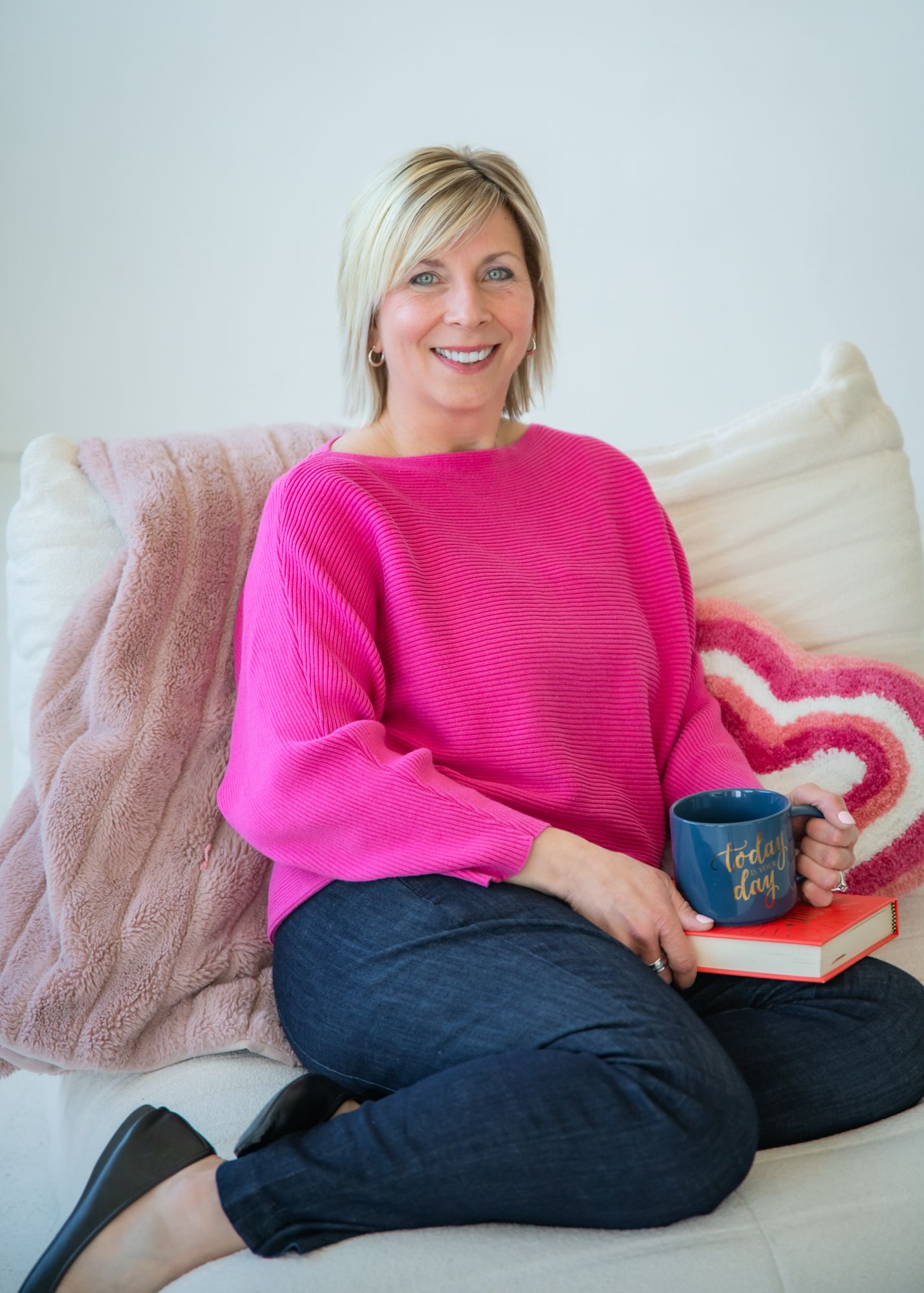 Smiling blonde woman in pink sweater sitting on white couch holding blue mug and book with heart pillow backdrop.