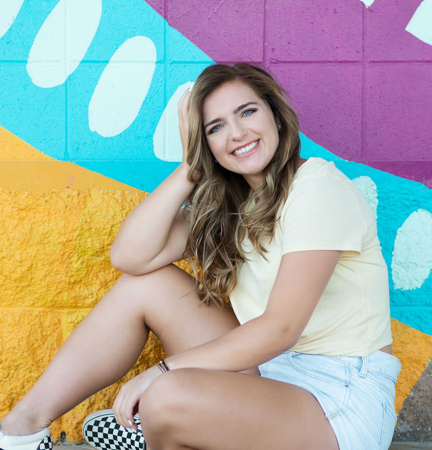 Smiling teenage girl in yellow top and checkered sneakers sitting against colorful mural wall.