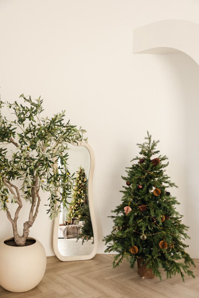 Small decorated Christmas tree beside an organic-shaped mirror and potted olive tree in a minimalist room with herringbone wood floors.