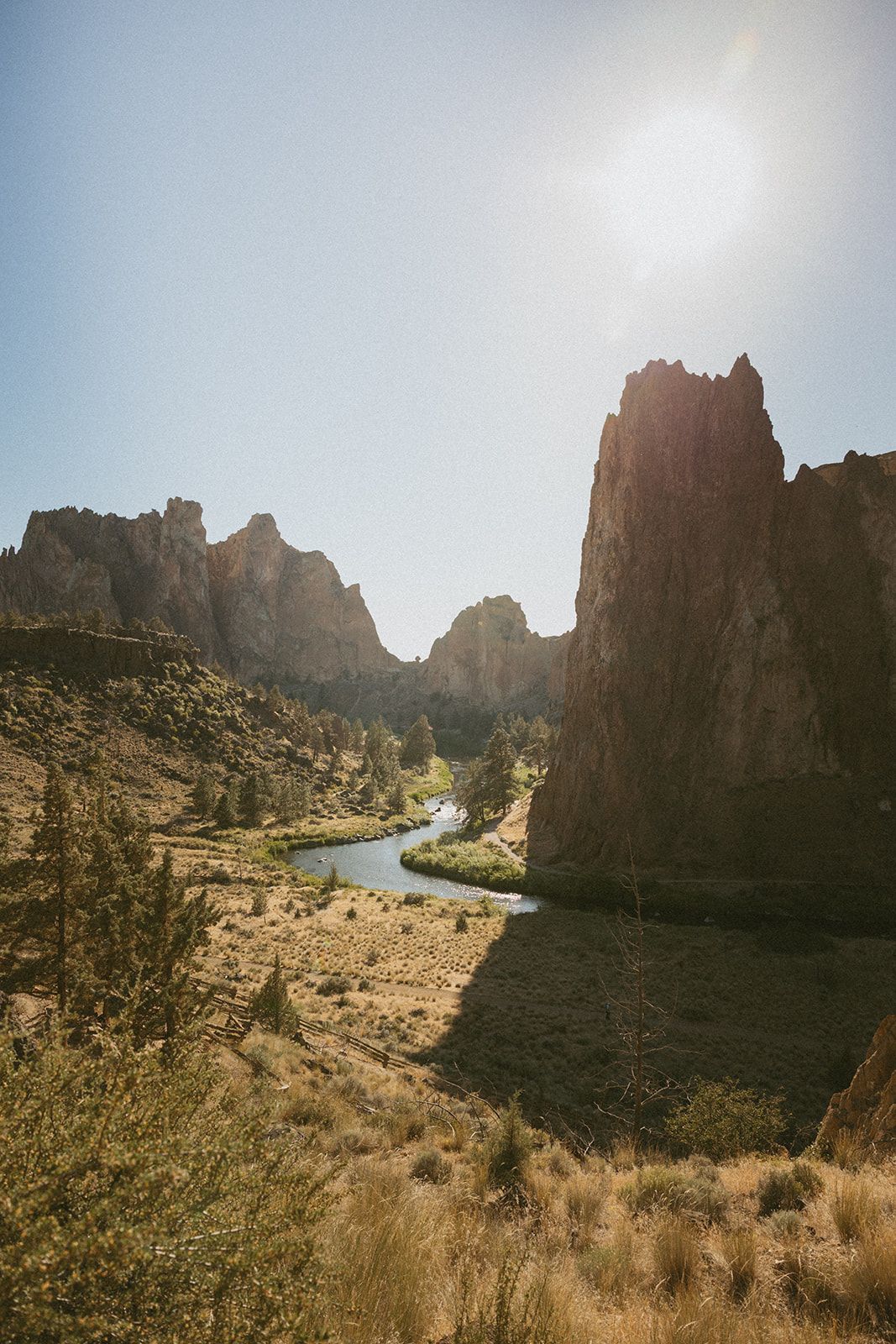 Golden sunlit rock cliffs reflect in a calm river lined with green grass and evergreen trees under a clear blue sky.