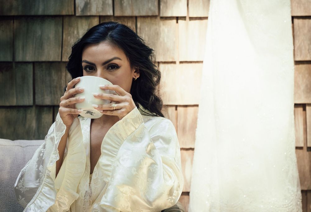 Bride in white satin robe sipping from a white mug near a wedding dress, with wooden shingle wall backdrop.