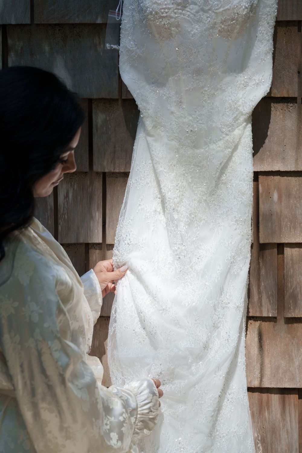 Bride in white robe examining her lace wedding dress hanging on a rustic wooden shingle wall.