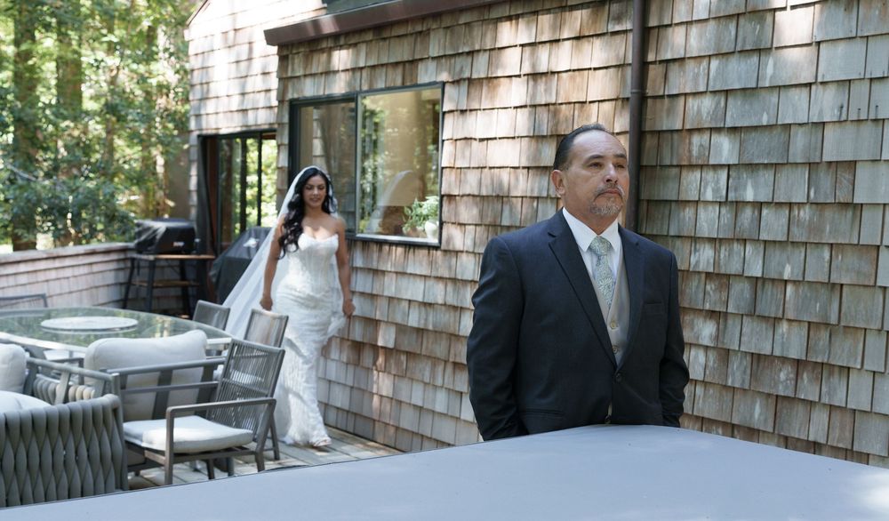 Bride in white wedding gown approaches man in suit outside rustic cedar-shingled venue with patio furniture.