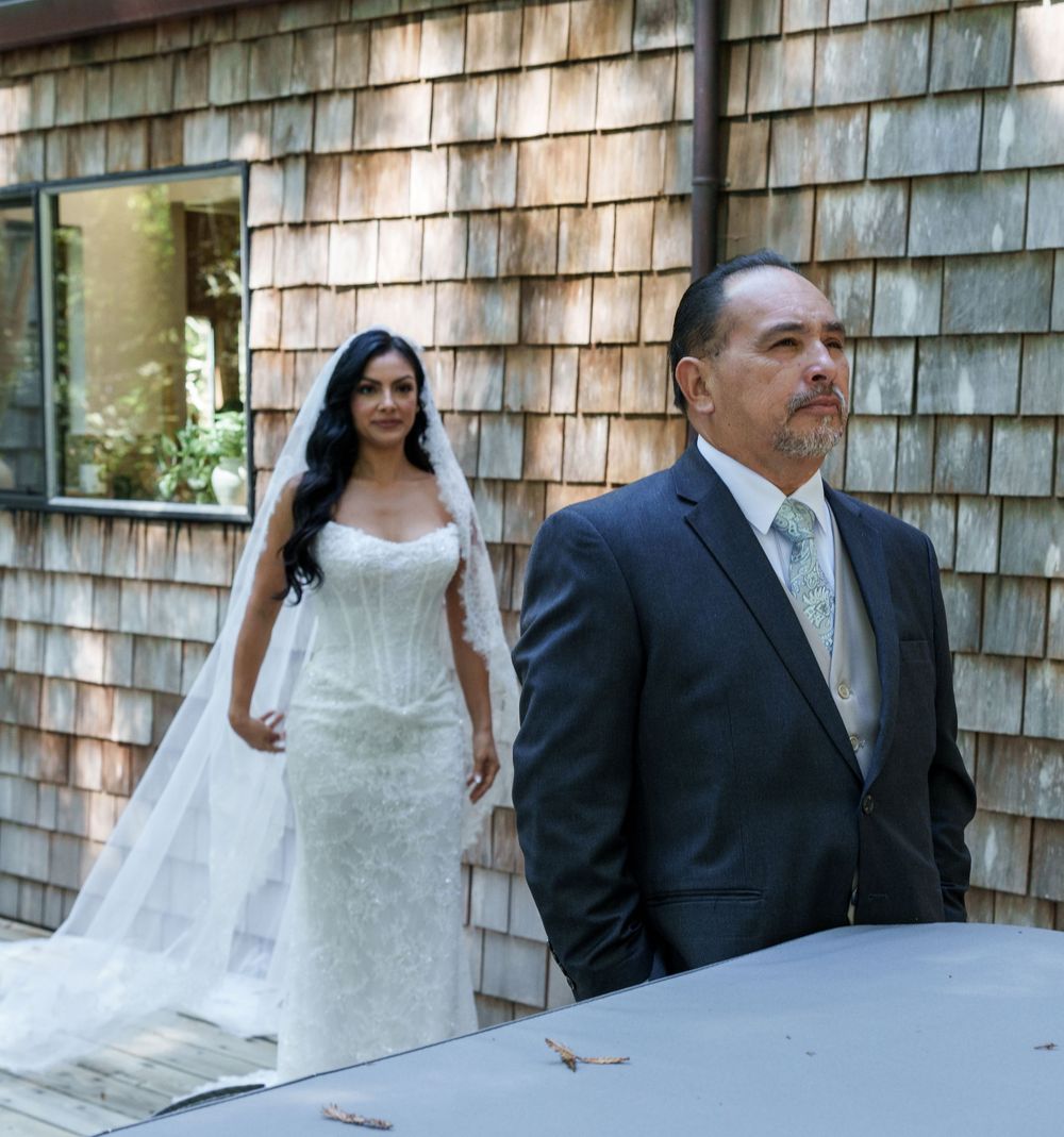 A bride in a white gown and veil stands behind a man in a gray suit during an outdoor wedding first look.