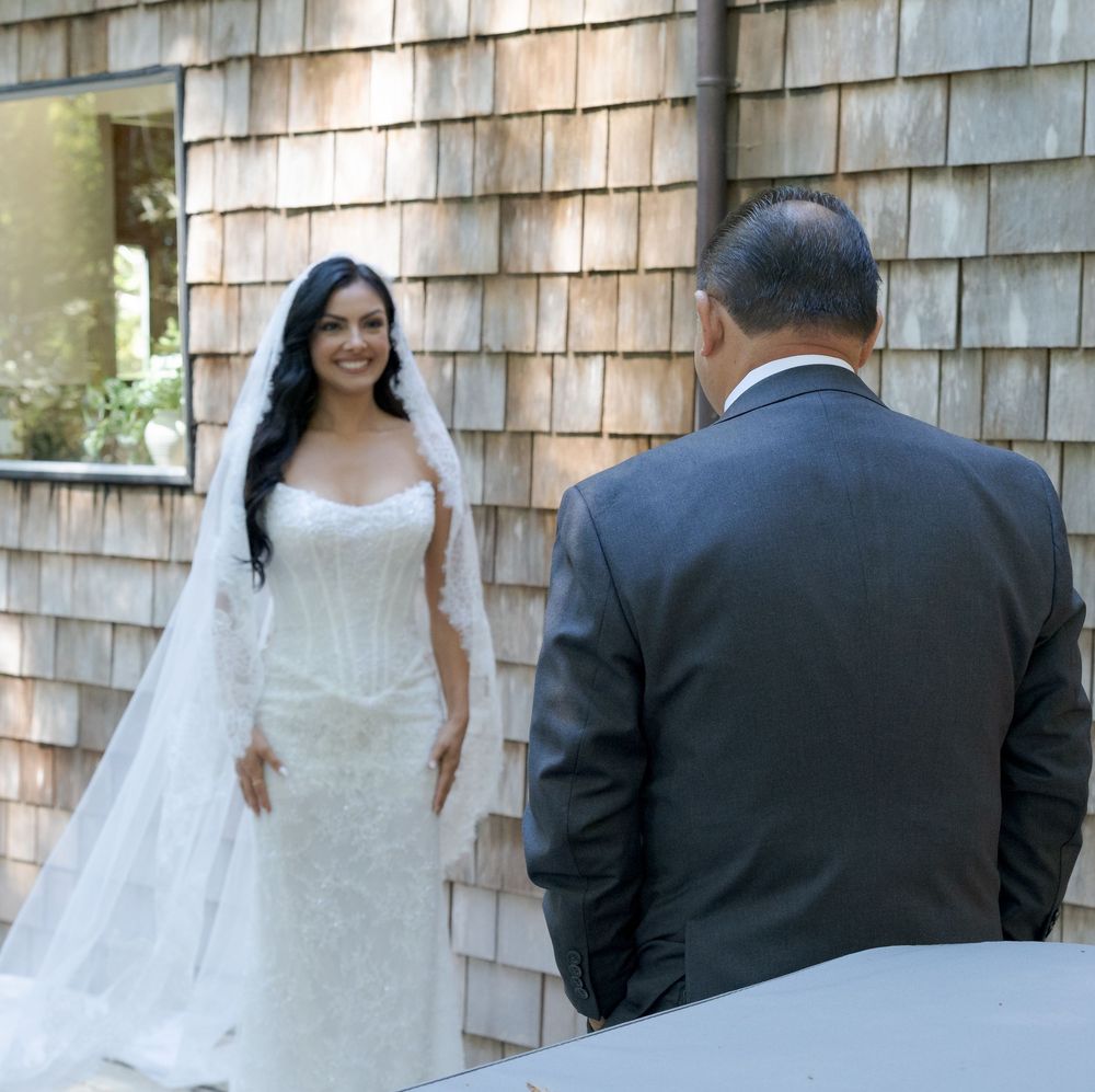 Bride in white lace gown and veil smiles during first look moment with groom in gray suit outdoors.