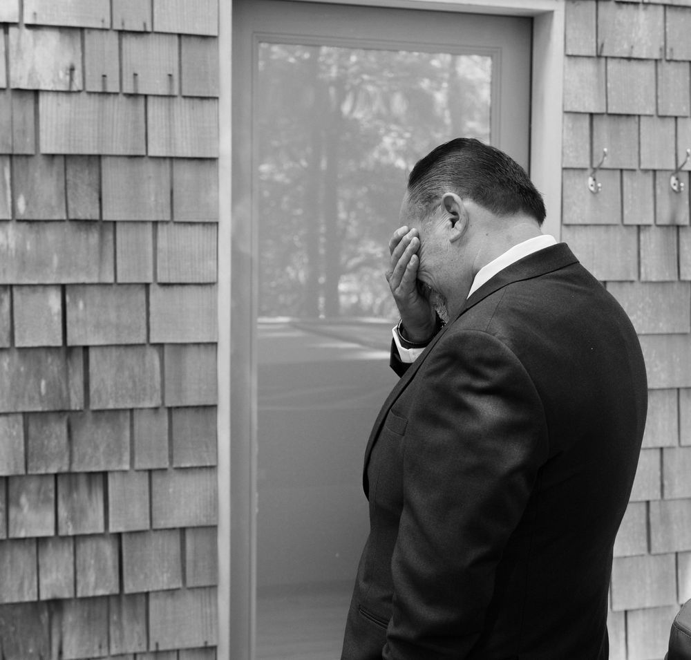 Black and white photo of a man in a suit covering his face with his hand outside a building with shingle siding.