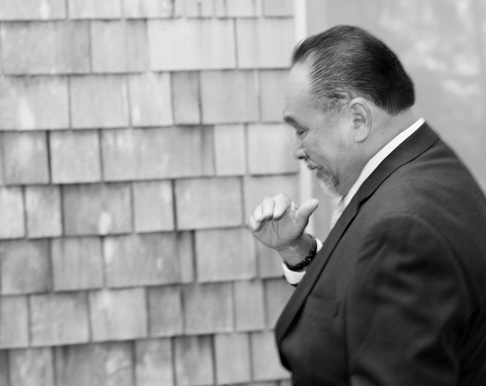 Man in dark suit standing in profile against a rustic wooden shingle wall in black and white photo.