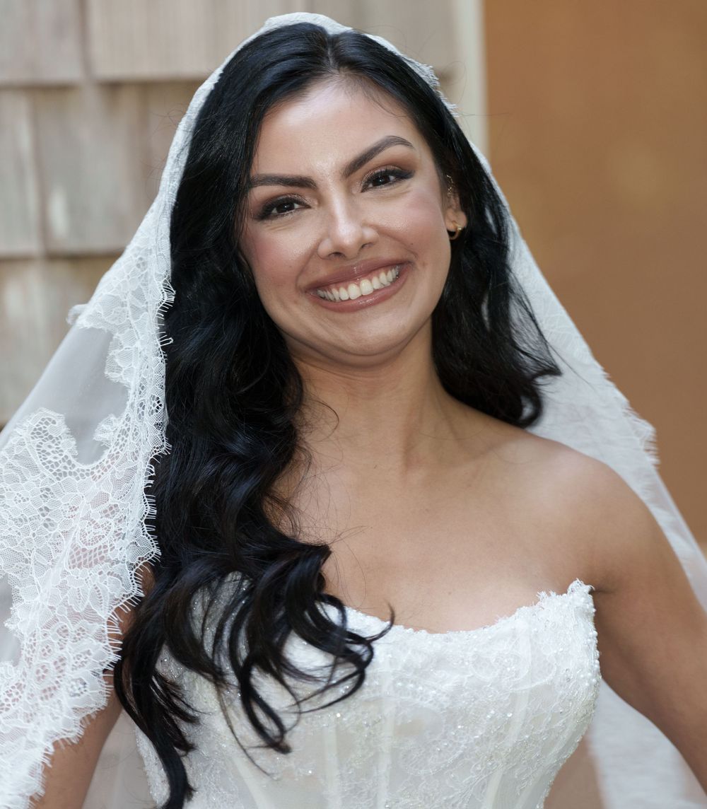 A smiling bride wearing a white strapless gown and lace veil with long dark wavy hair on her wedding day.