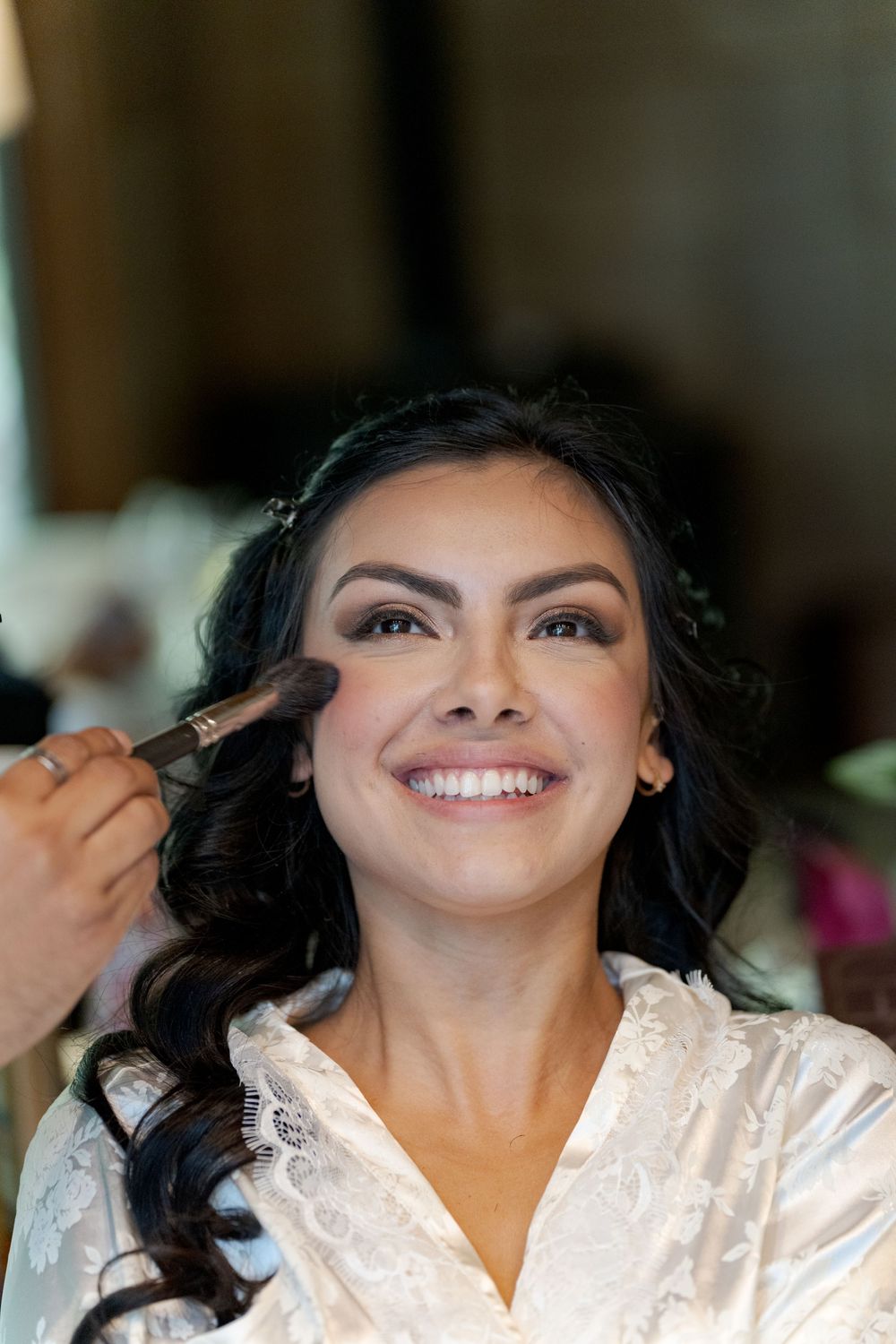 Smiling bride in white lace robe getting makeup applied with a brush on her cheek during bridal preparation.