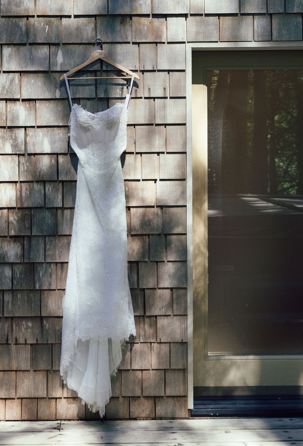 White lace wedding dress hanging on wooden hanger against rustic cedar shake siding near a door.