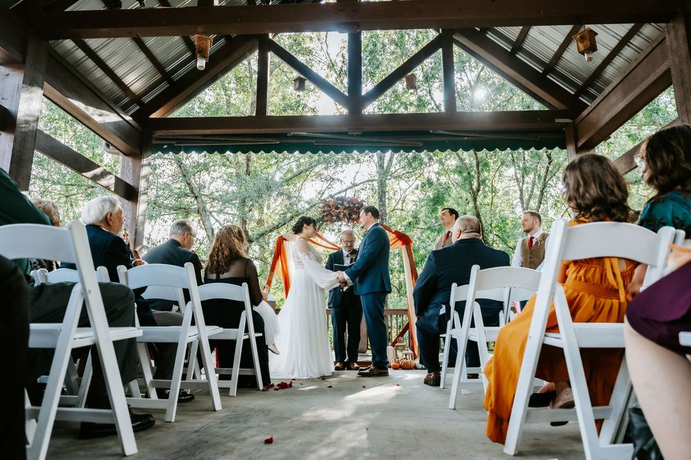 Outdoor wedding ceremony under a wooden pavilion with white chairs and colorful fall decorations.