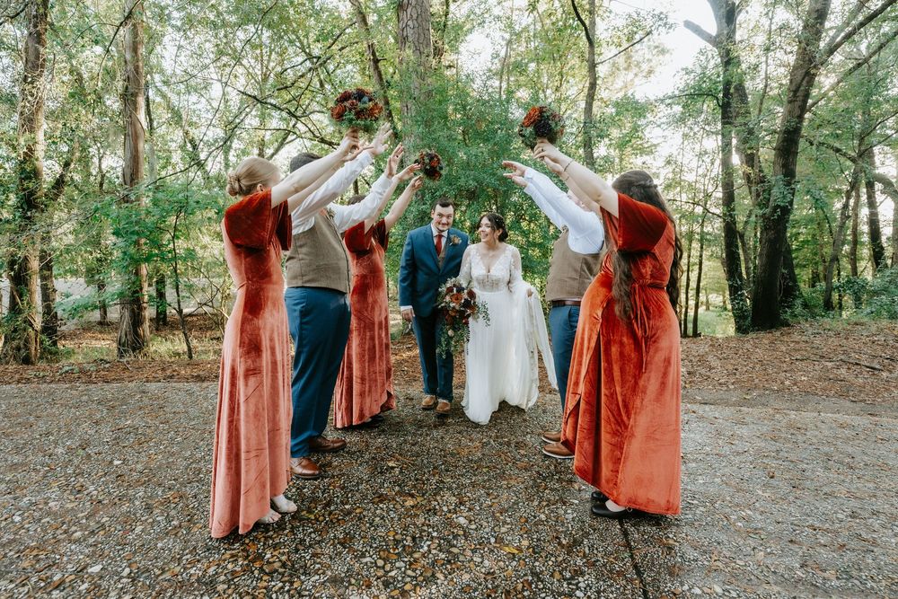 Wedding party in forest, bridesmaids in rust velvet dresses raising bouquets to form arch over bride and groom.