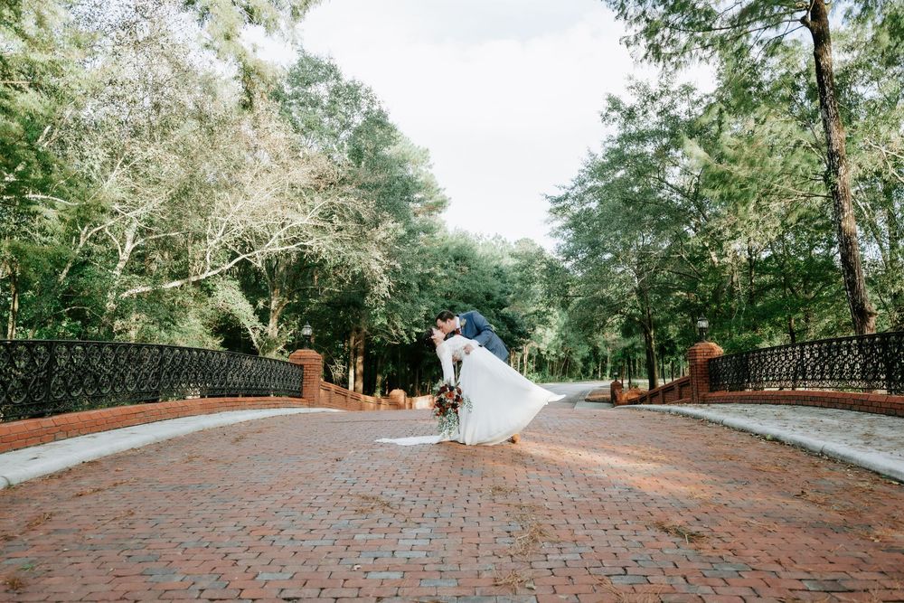 Bride and groom share a romantic dip kiss on a brick pathway surrounded by lush green trees at an outdoor wedding venue.